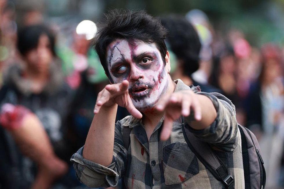 A person in zombie makeup with milky eye, bloody shirt, and outstretched arms at zombie walk.