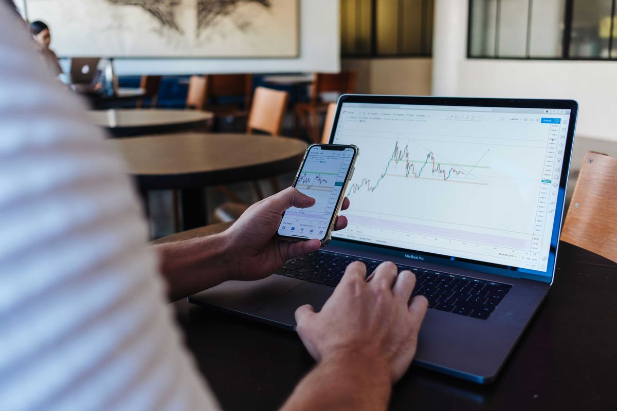 A person's hands operating a laptop and smartphone, both displaying financial trading charts.
