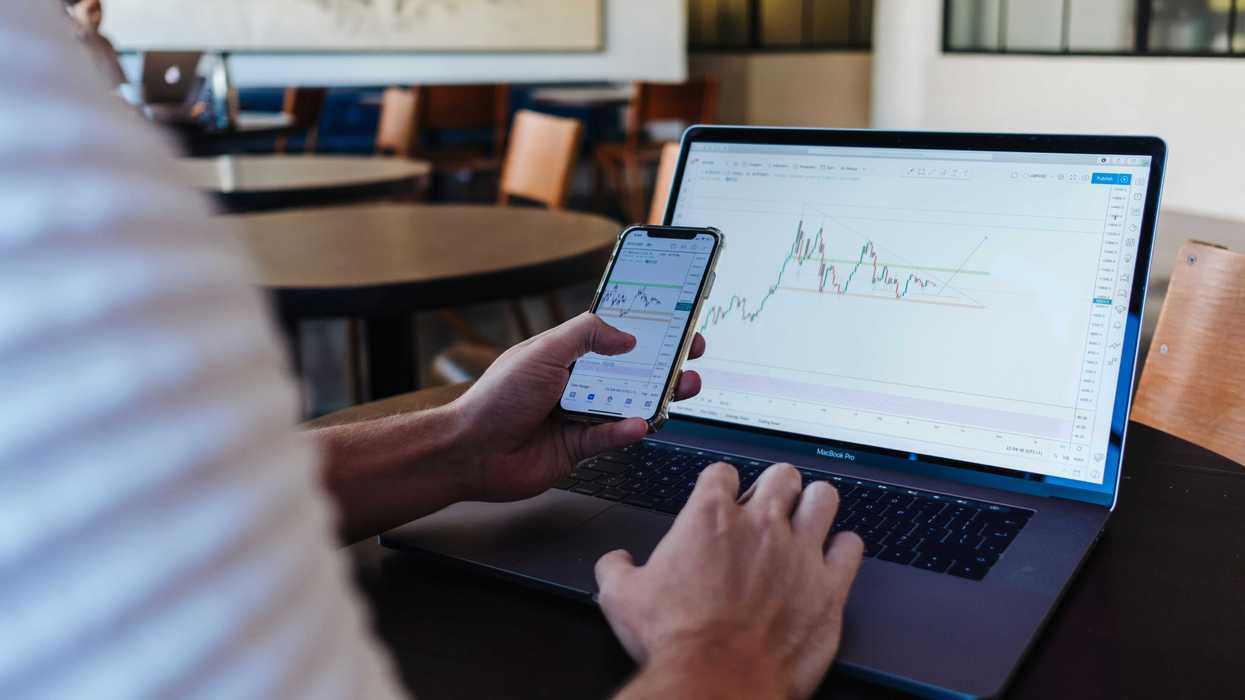 A person's hands operating a laptop and smartphone, both displaying financial trading charts.