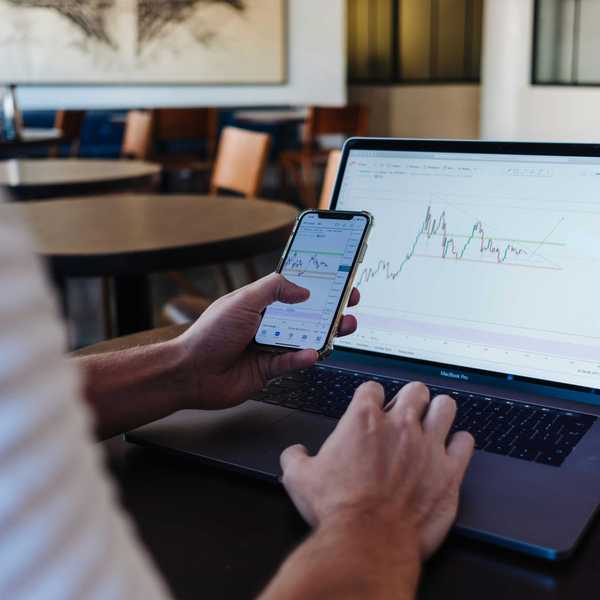 A person's hands operating a laptop and smartphone, both displaying financial trading charts.