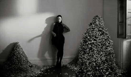 Charl XCX smokes a cigarette while walking among big piles of cigarettes