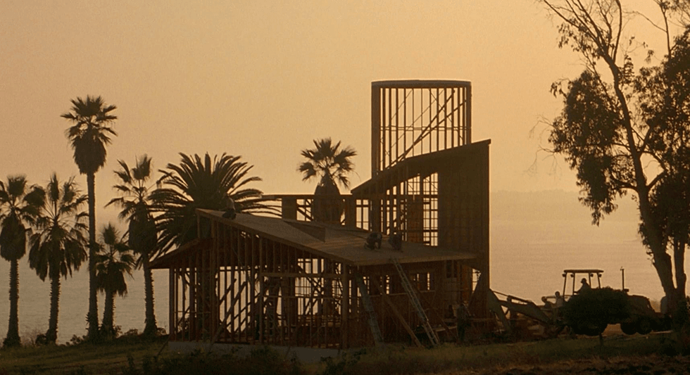 Construction site silhouette at sunset: wood-framed house, palm trees, ocean view, and a tractor
