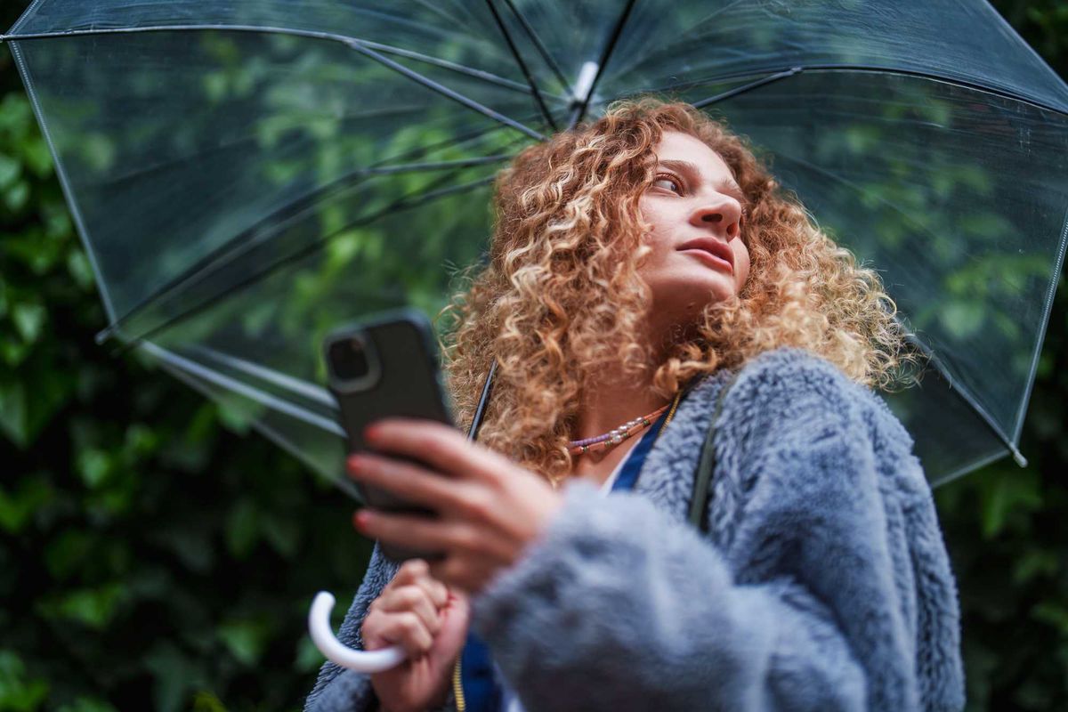 Curly-haired woman under clear umbrella holds phone, looking upwards