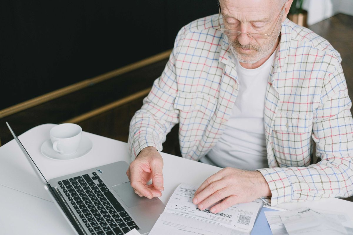 Elderly man in glasses reviewing bills at a table with a laptop and coffee cup