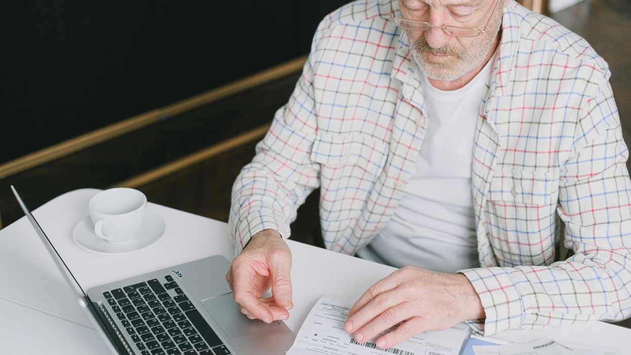 Elderly man in glasses reviewing bills at a table with a laptop and coffee cup