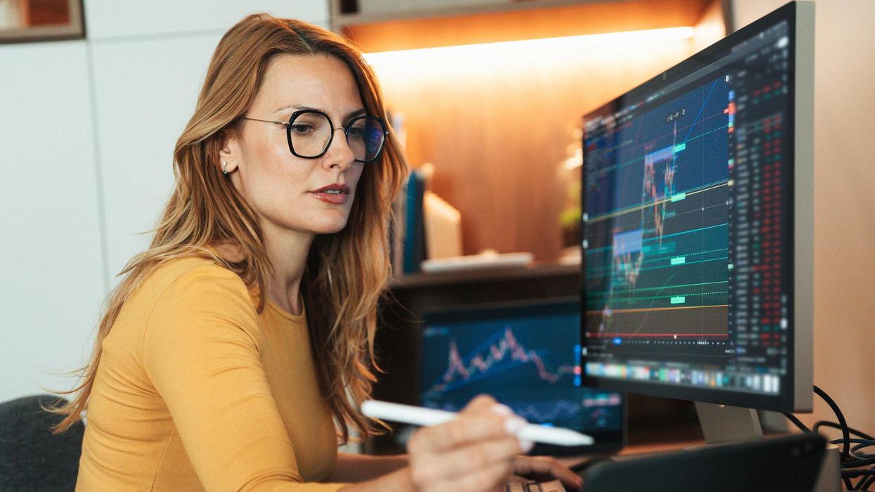 Female trader with glasses monitors financial charts on dual screens, holding a digital pen.