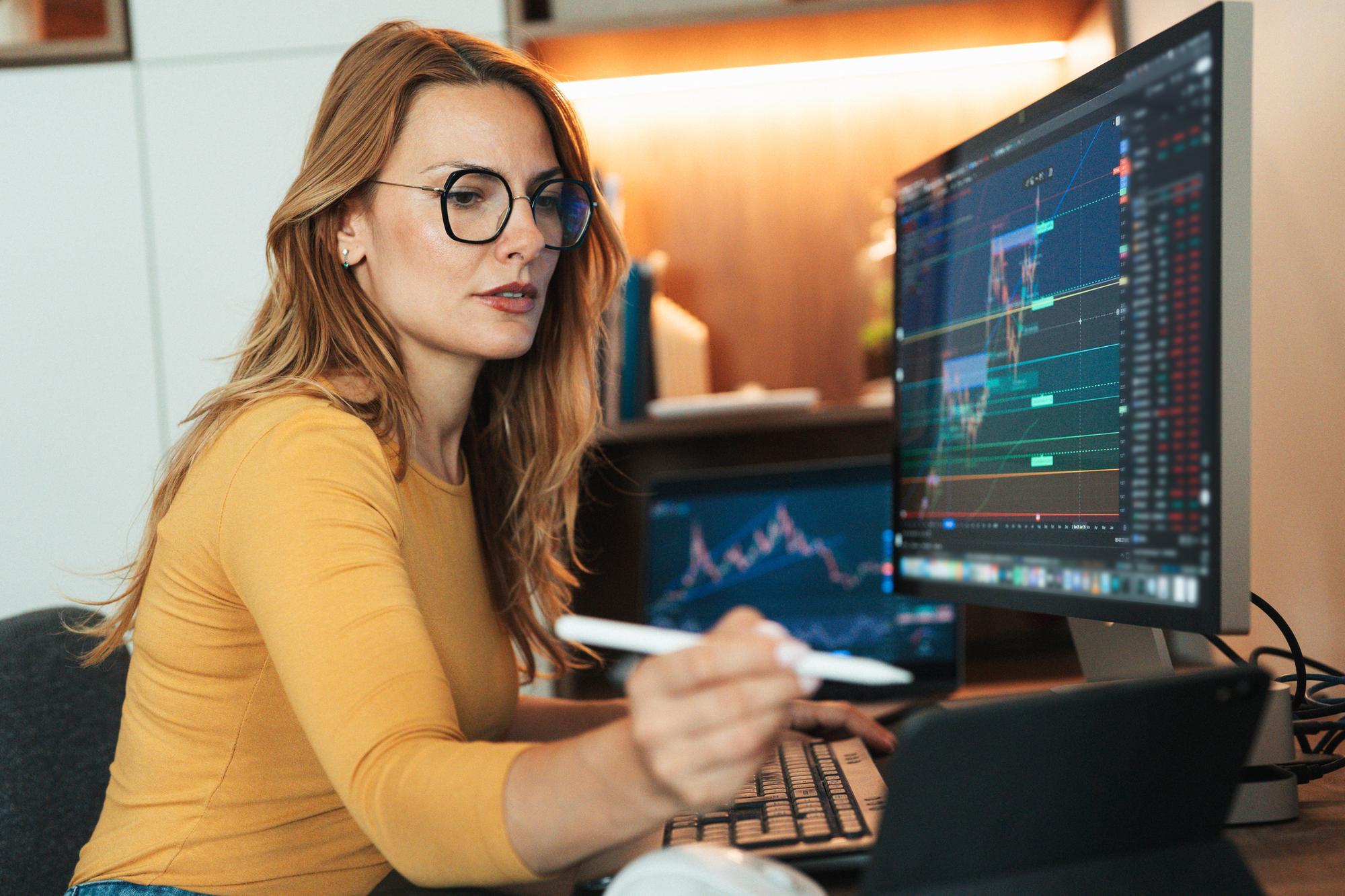 Female trader with glasses monitors financial charts on dual screens, holding a digital pen.