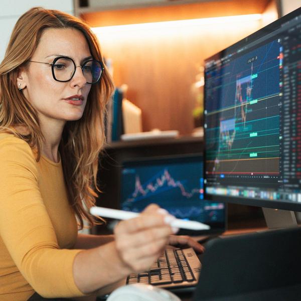 Female trader with glasses monitors financial charts on dual screens, holding a digital pen.