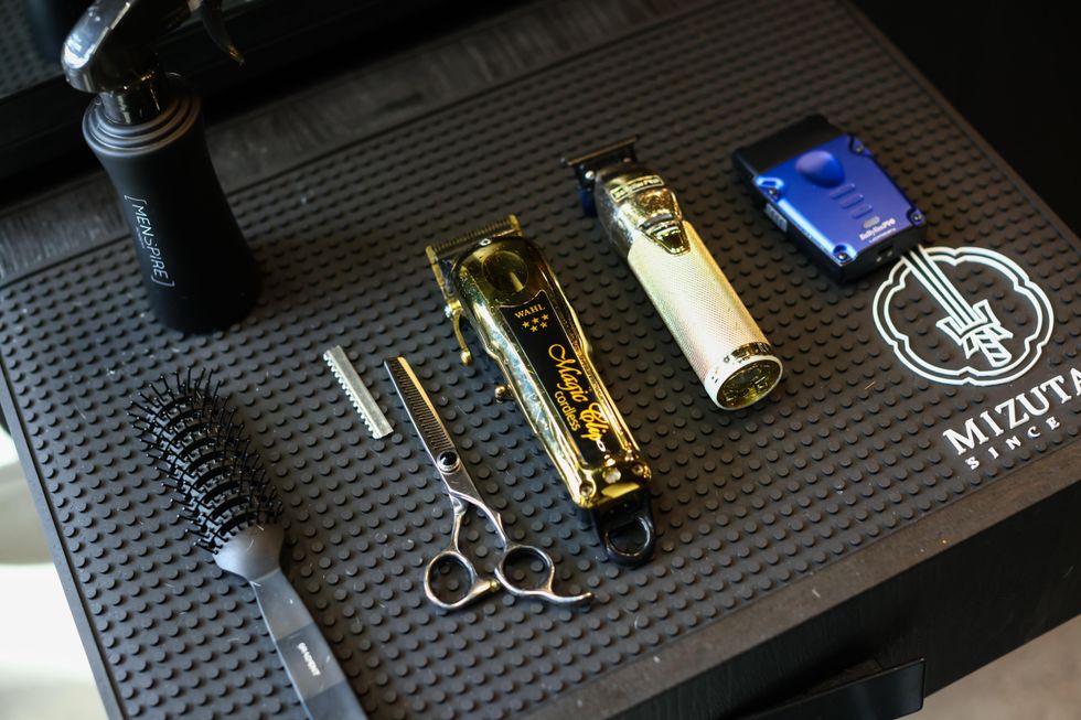 Gold clippers, blue shaver, thinning shears, brush, and spray bottle on a barber's black textured mat