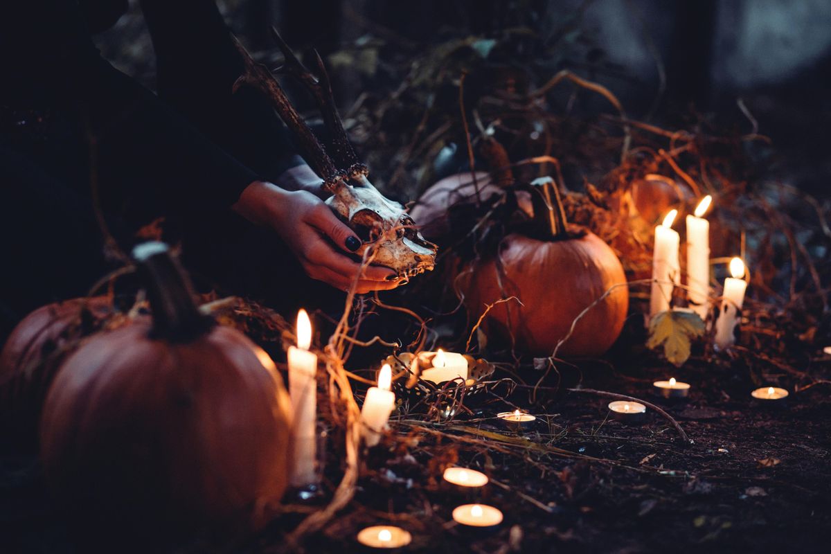 Hands holding an antlered deer skull, surrounded by pumpkins and many lit candles in the dark.