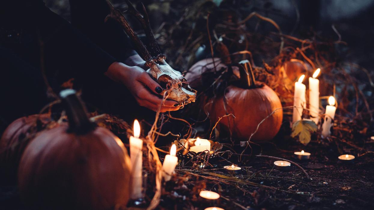 Hands holding an antlered deer skull, surrounded by pumpkins and many lit candles in the dark.