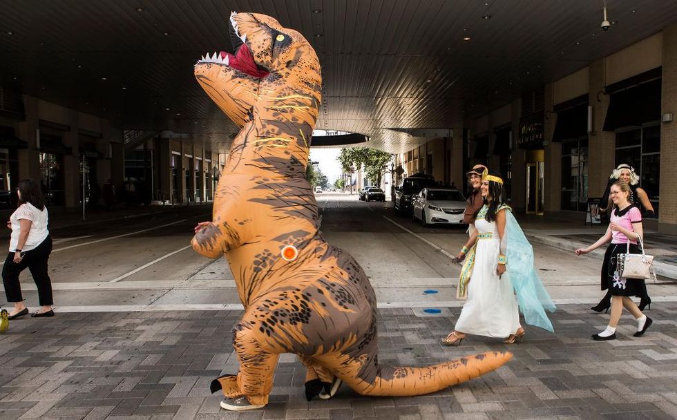 Person in inflatable T-Rex costume walks with costumed people on a city street.