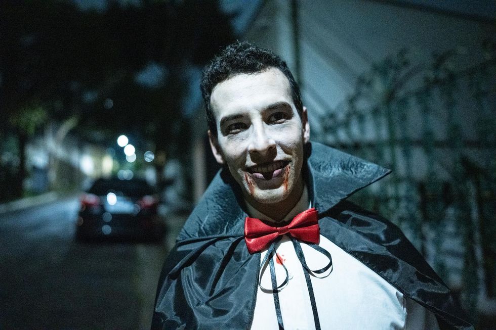 Smiling man in vampire costume with white face makeup, red bowtie, and blood on his mouth.