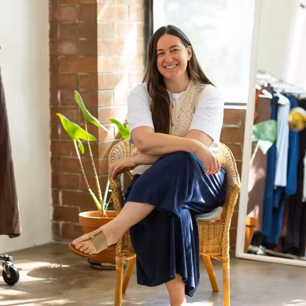 Smiling woman in white shirt and vest sits in a wicker chair in a clothing store