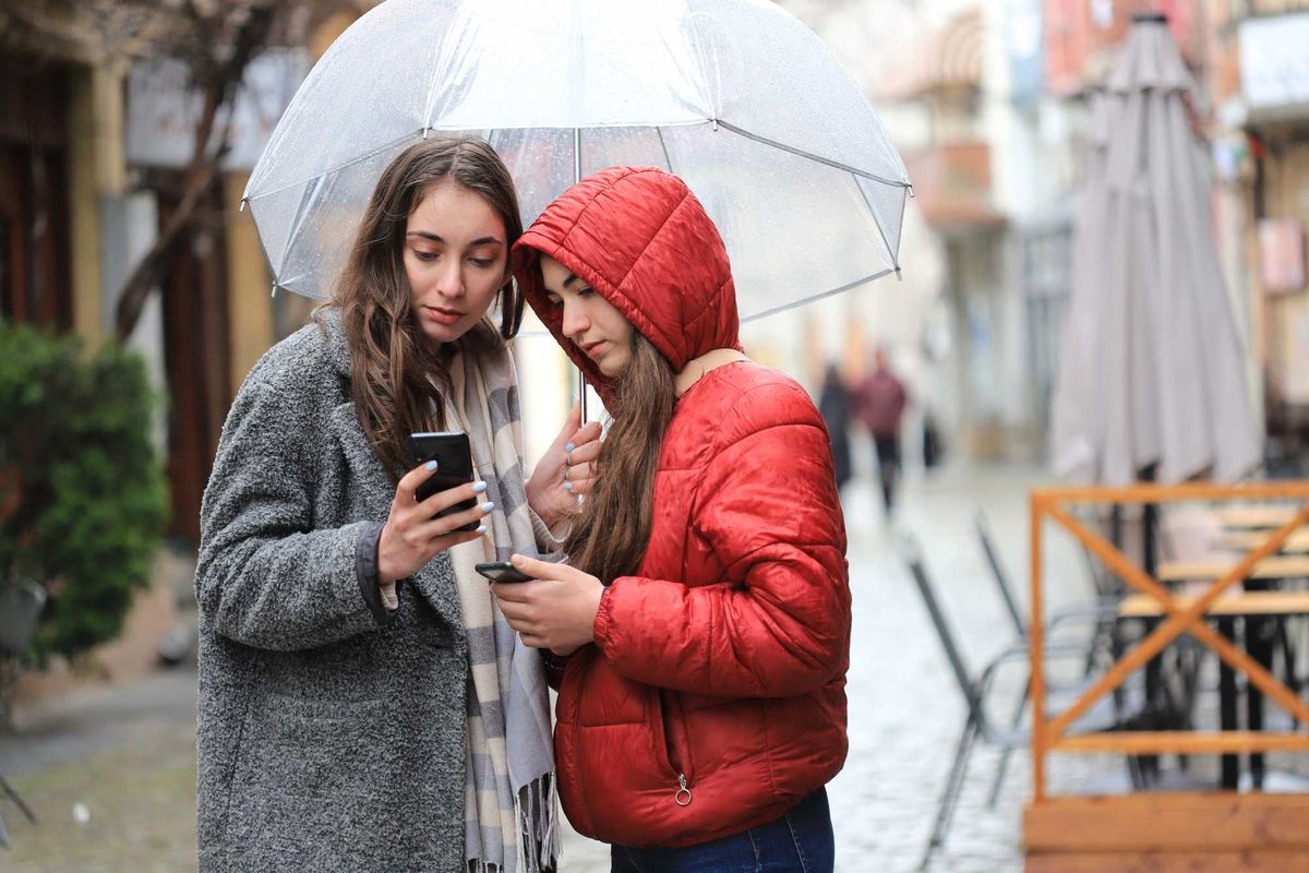 Two women under an umbrella check their investments on their phones
