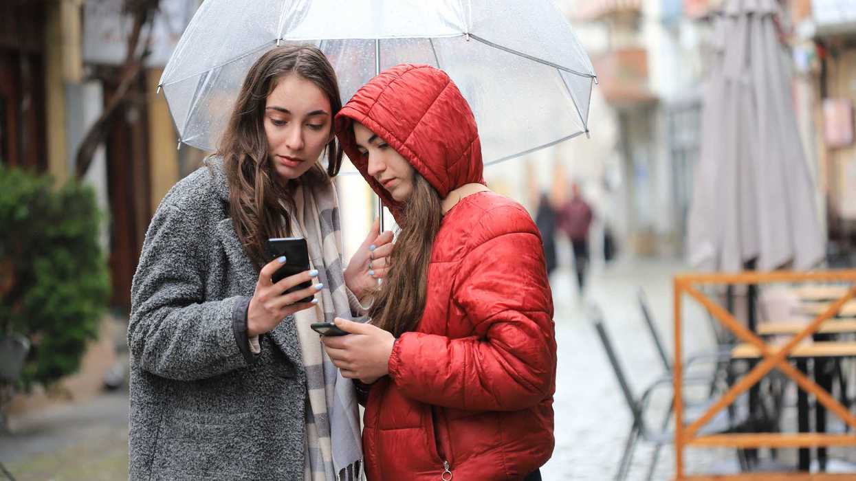 Two women under an umbrella check their investments on their phones