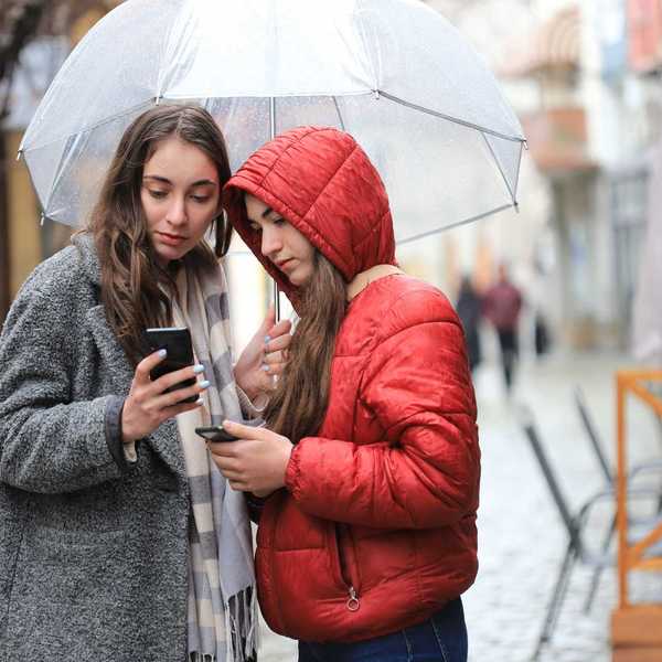 Two women under an umbrella check their investments on their phones